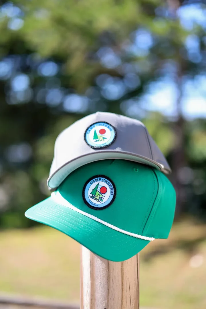 Close-up of a green rope hat and grey cap stacked on a post, featuring custom embroidered Camp Cheerio patches.