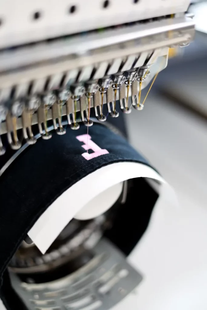 Embroidery machine stitching a pink letter onto a black and white visor